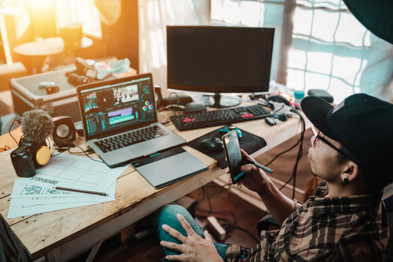 The content creator using mobile working with the laptop editing video footage in the house studio