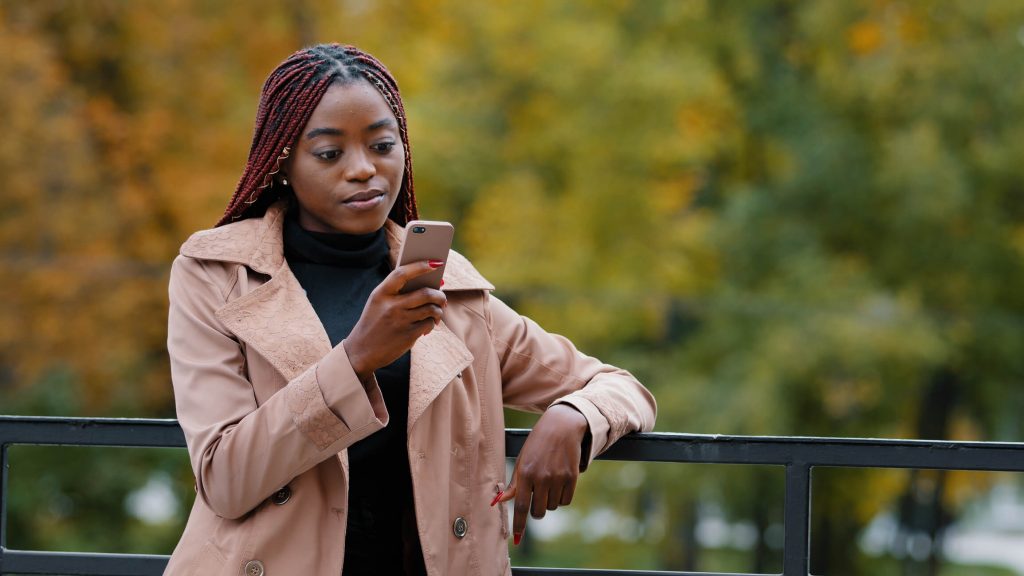 Young serious female standing in autumn park holding mobile phone browsing email remotely communicating